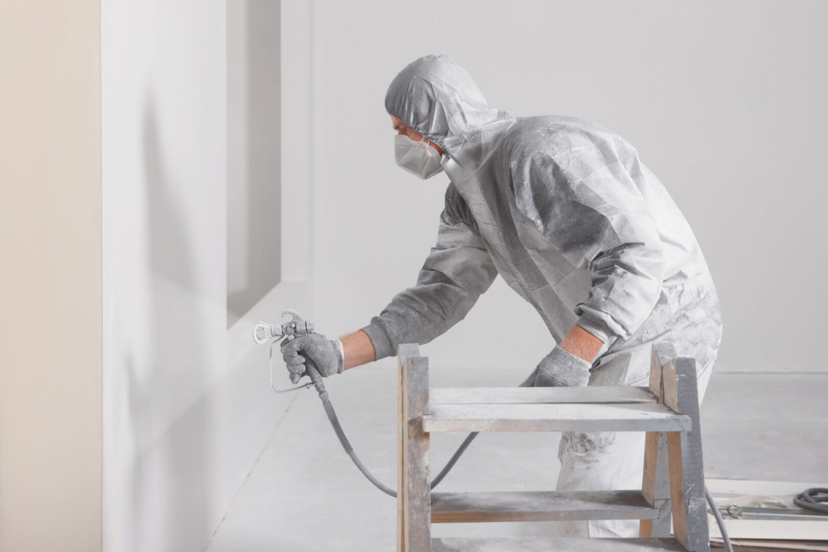 Decorator in protective overalls painting wall with spray gun indoors